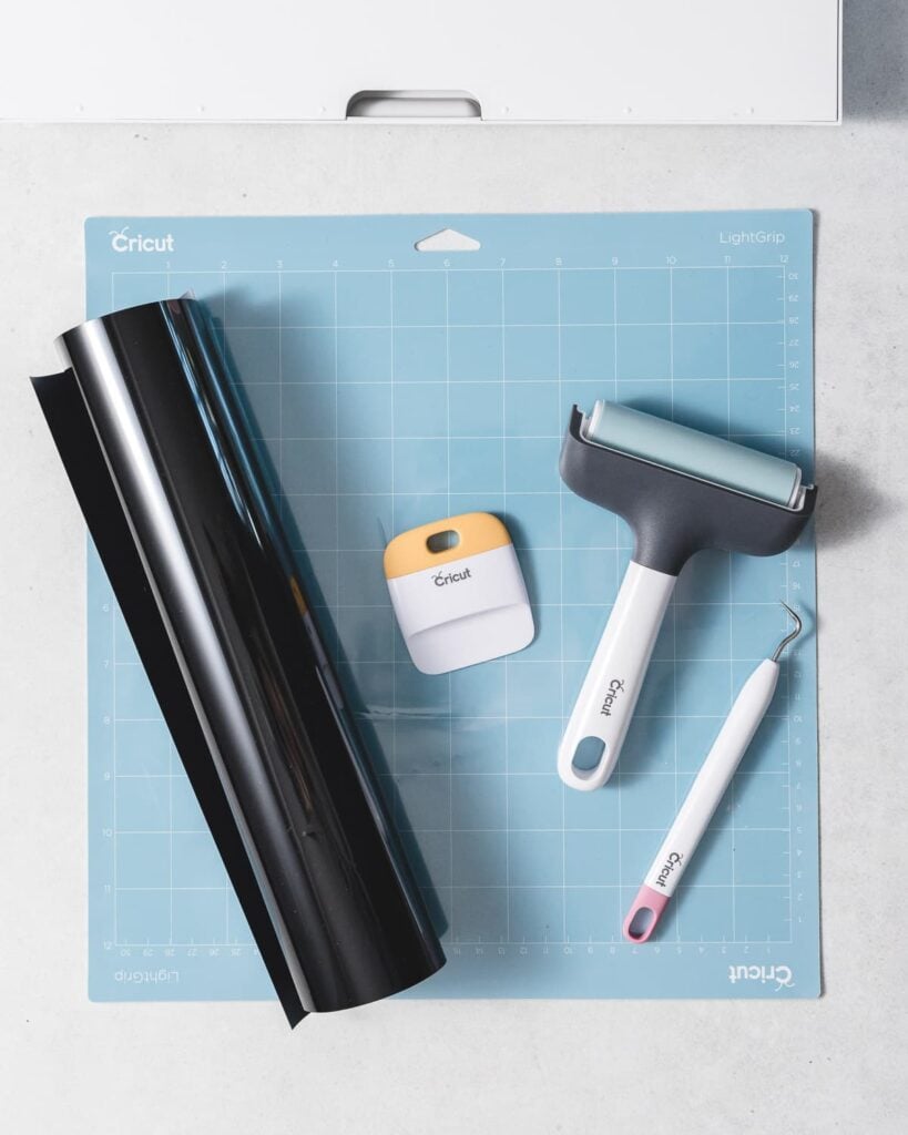 a roll of black vinyl, a scraper tools, a brayer, and a weeding tool on top of a blue cricut machine mat