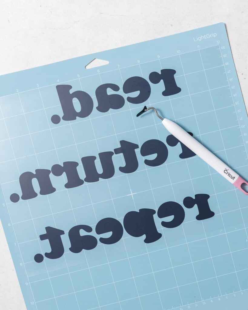 a weeding tool on top of a text-based design in black vinyl on a blue cutting mat.