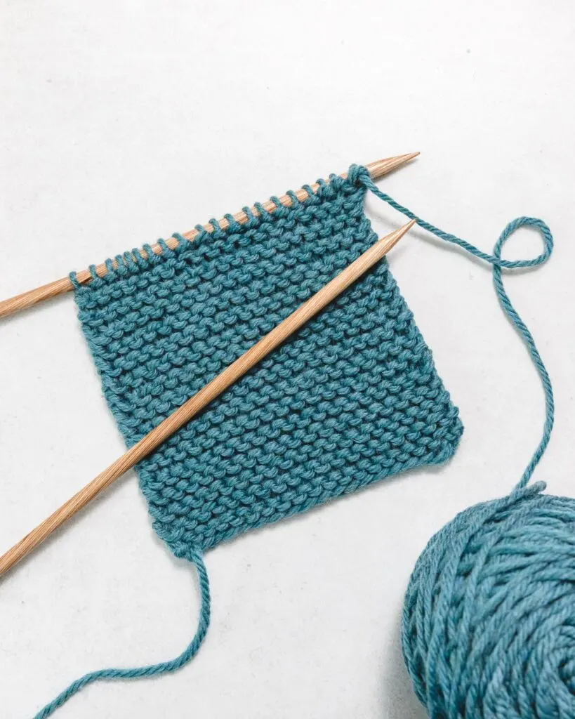 a swatch of garter stitch fabric in blue yarn with brown wooden knitting needles and a ball of yarn